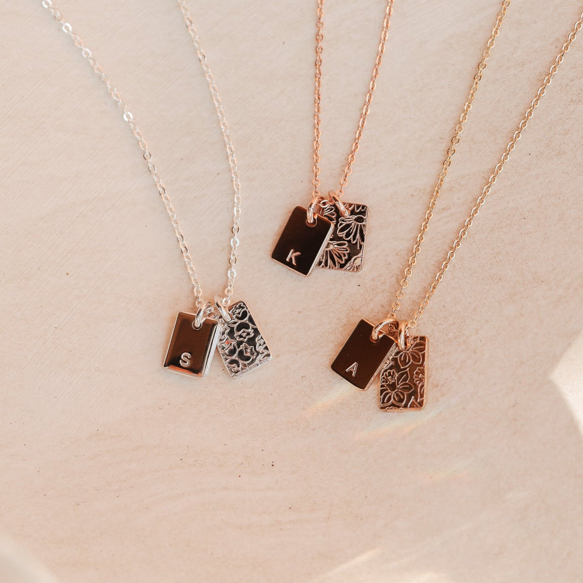 Three necklaces with small rectangular initial pendants on a light pink background