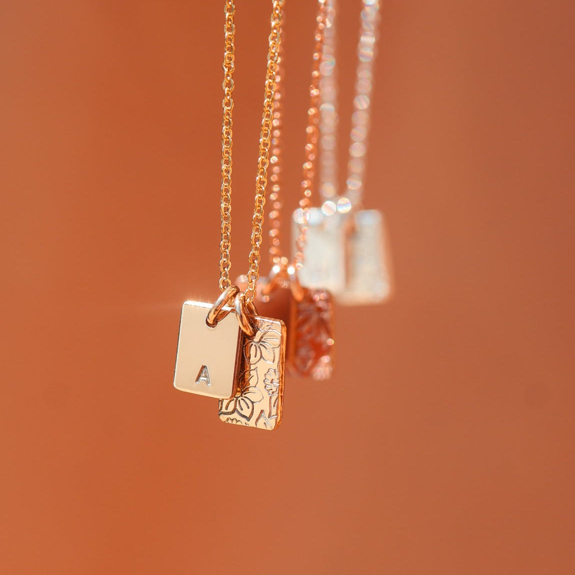 Three necklaces with initials and scattered birth flowers on square pendants on a warm brown background