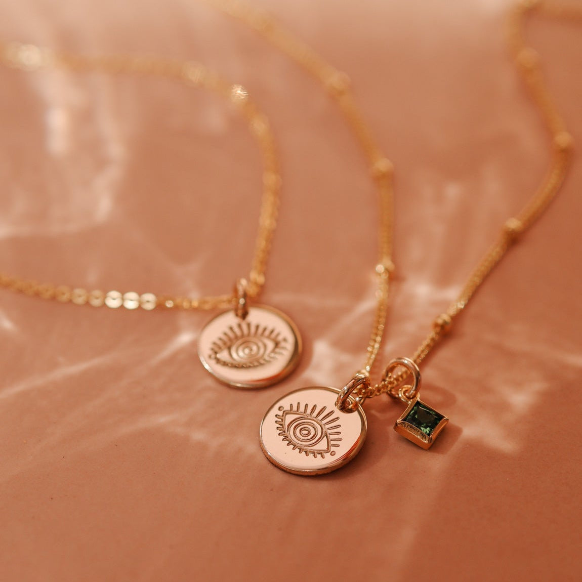 Two delicate gold satelite chain necklaces with small evil eye pendants and a small square green birthstone accent. Brisbane personalised jewellery