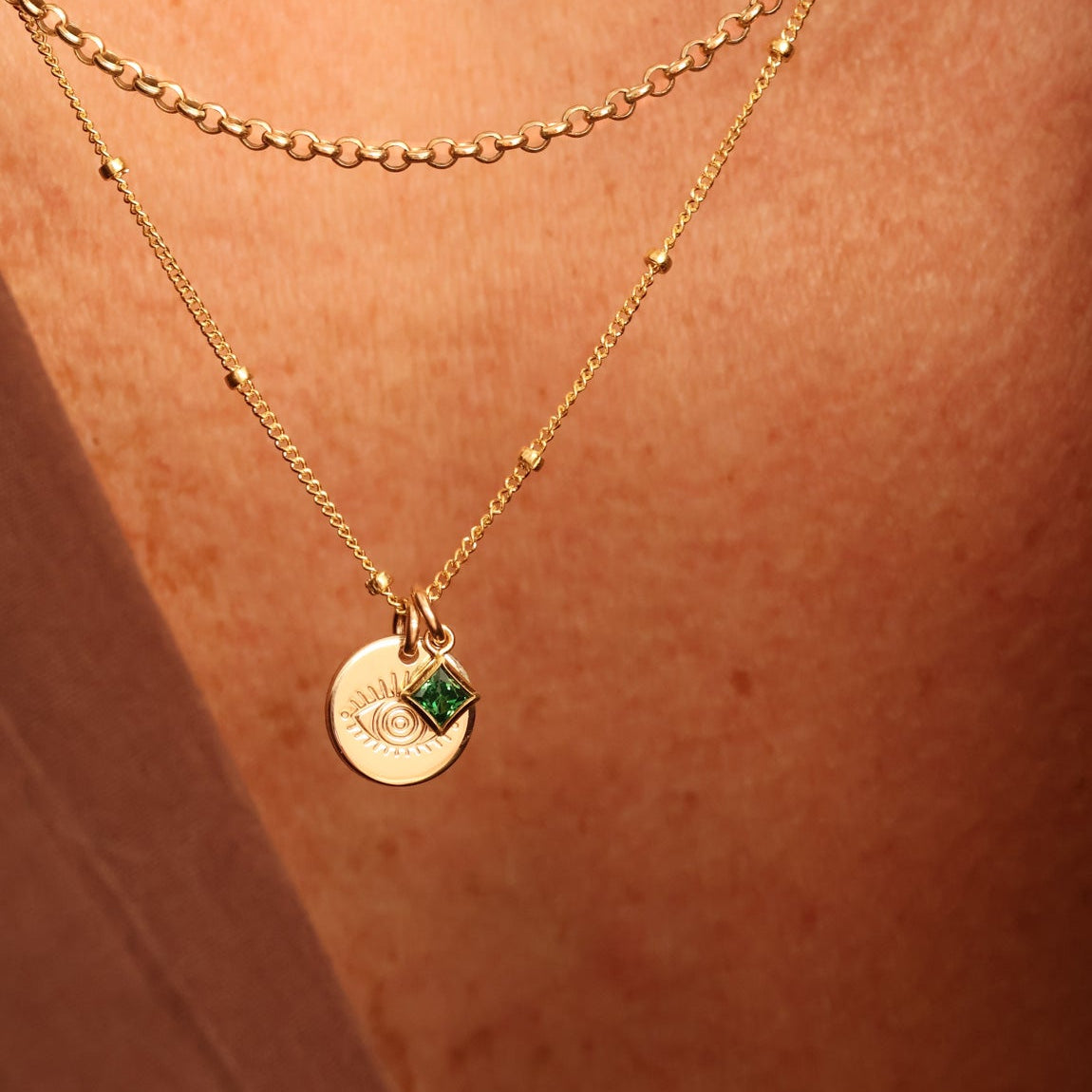 A close-up of a person's chest wearing a delicate gold satelite chain necklace with small evil eye pendant and a small square green birthstone accent. Sturdy rolo layer chain. Brisbane personalised jewellery